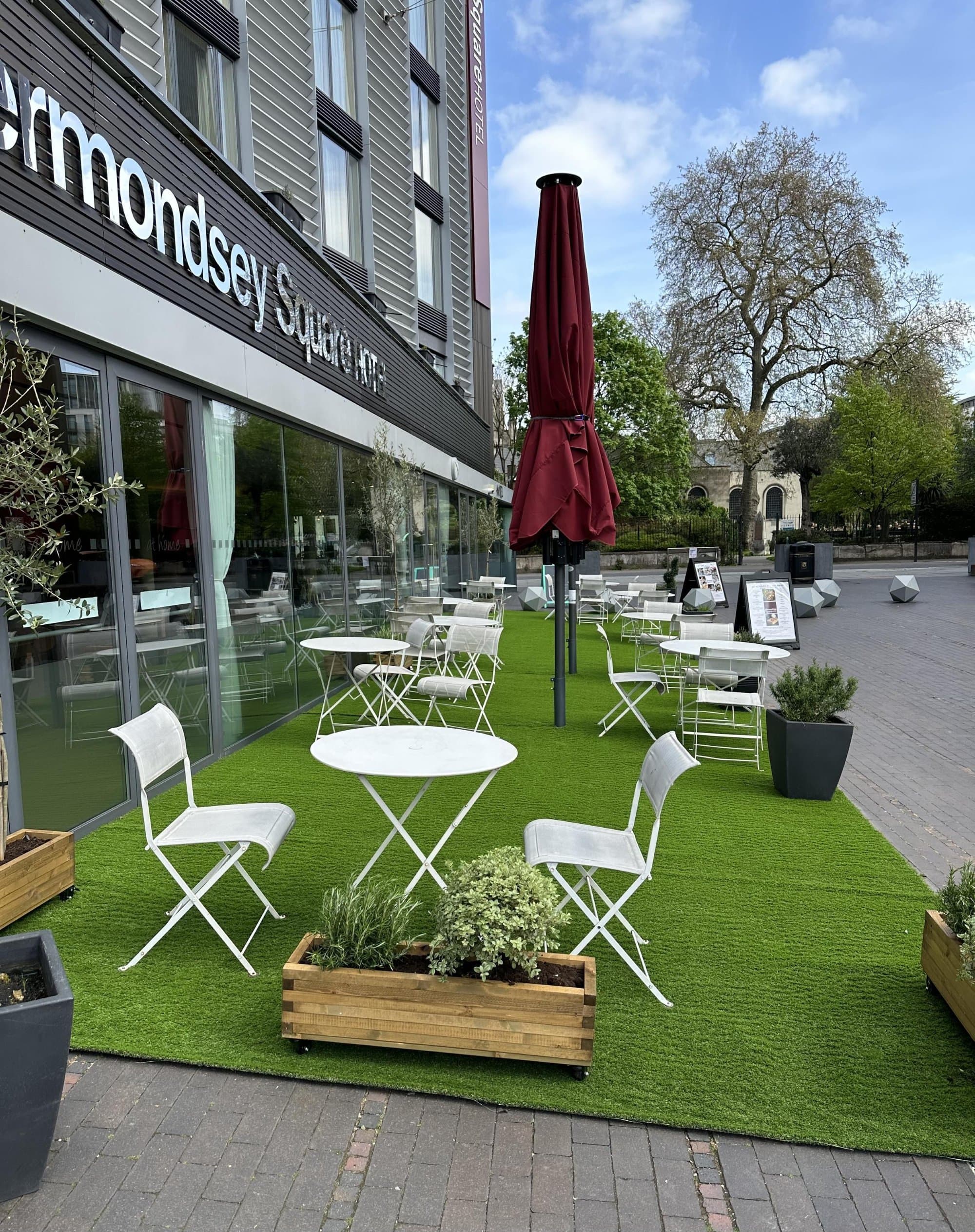 A white café table and chairs set up on freshly laid artificial lawn outside The Bermondsey Square Hotel, with a wooden planter of white flowers in the foreground and a red umbrella furled behind